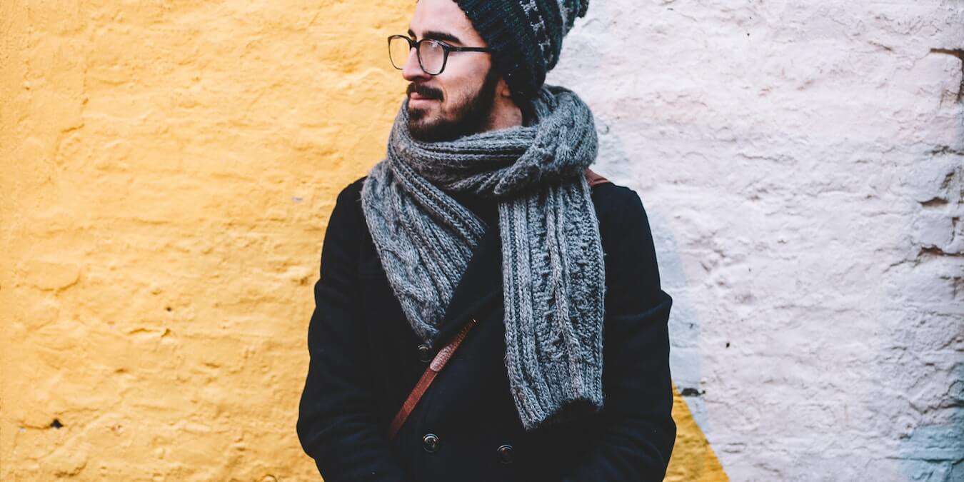 Man in glasses and knit hat wears gray cable knit scarf in front of yellow and white painted wall.